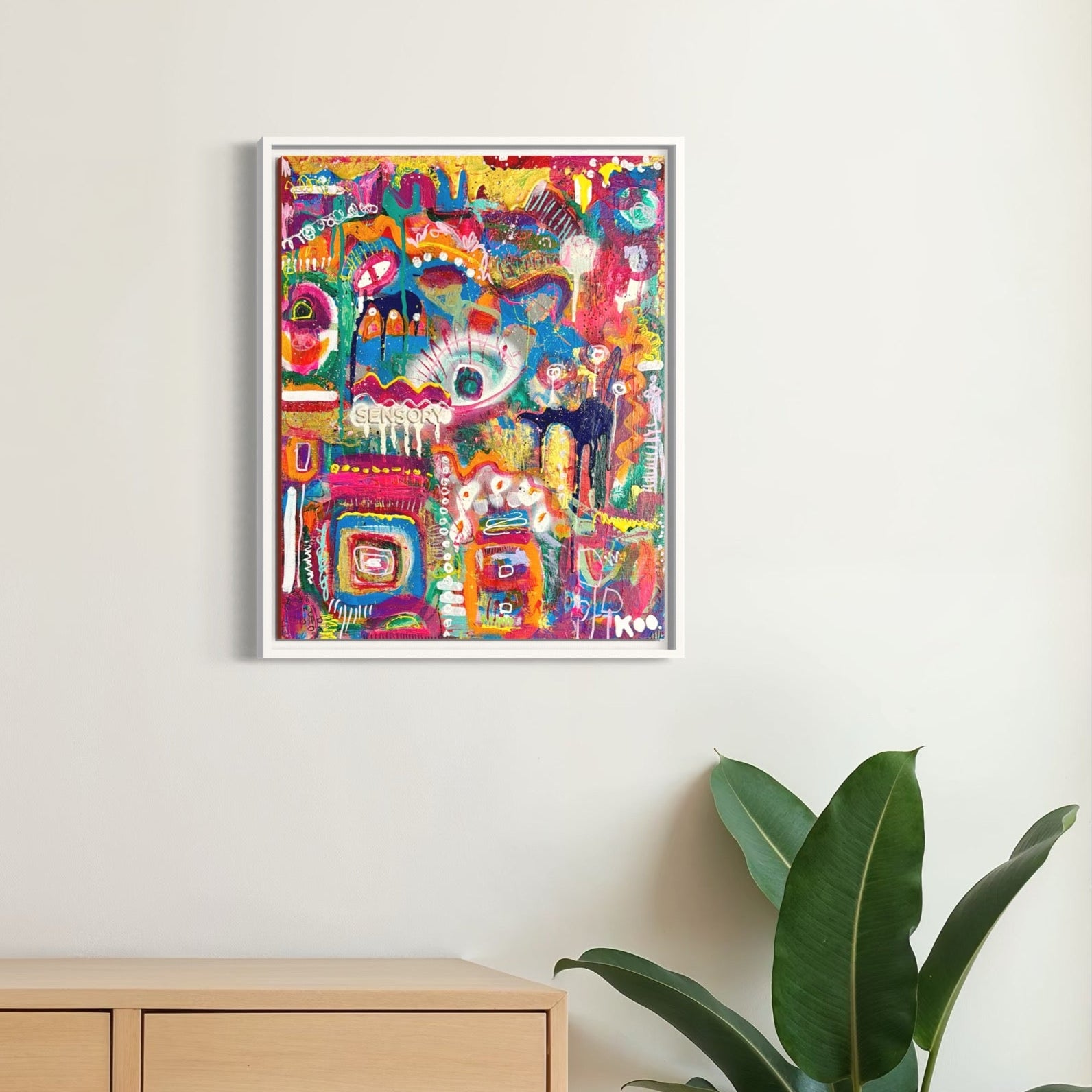 Colorful abstract art piece on a wall above a wooden desk with a lamp and plants.