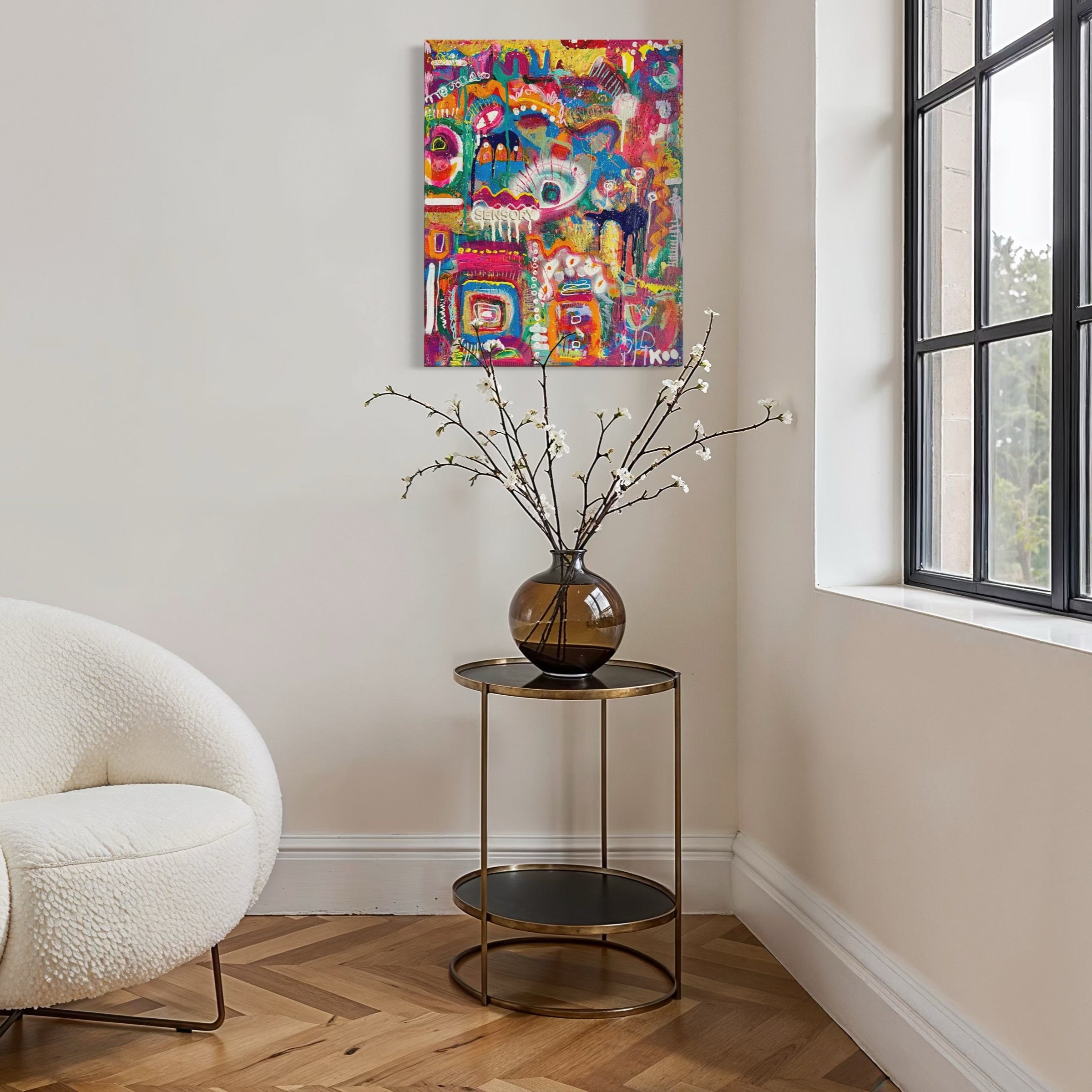 White armchair in a room with a colorful abstract painting on the wall and a small table with a vase and branches.